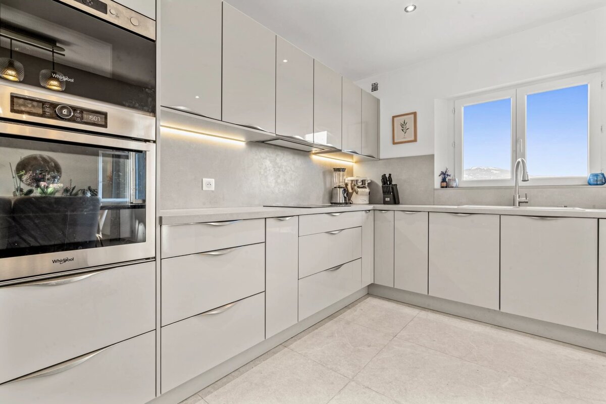 A modern, bright kitchen features light grey cabinets, built-in appliances, under-cabinet lighting, and a large window with a scenic view.