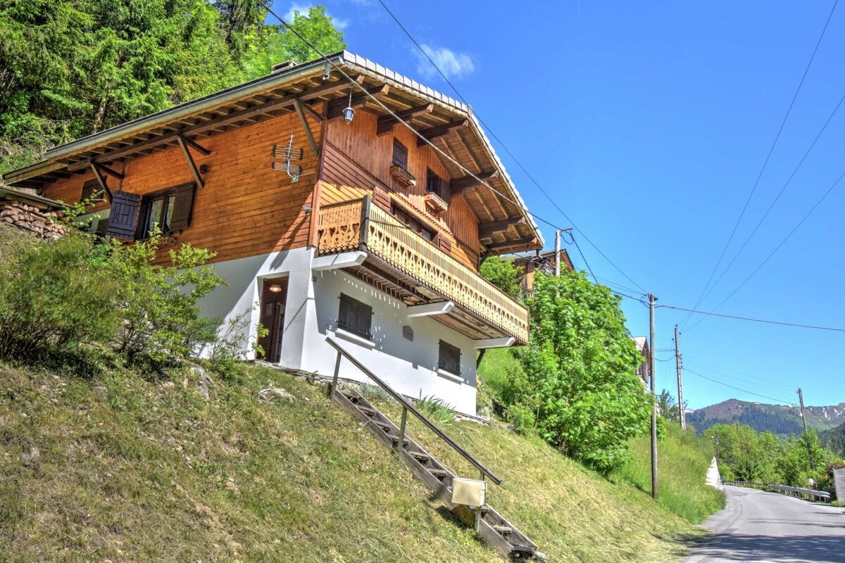 A charming wooden chalet with a balcony sits on a steep green hill, nestled in a lush, mountainous landscape under a bright blue sky.