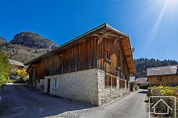A large rustic wooden and stone building in a sunny, mountainous setting with a clear blue sky and a road.
