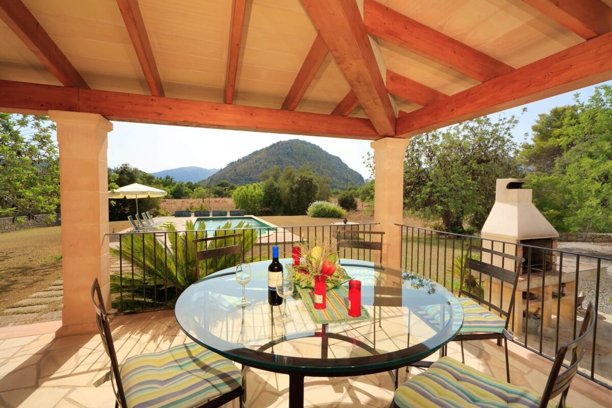 A covered patio with a glass table, wine, and fruit, overlooks a swimming pool, lush green landscape, distant mountains, and a BBQ grill.