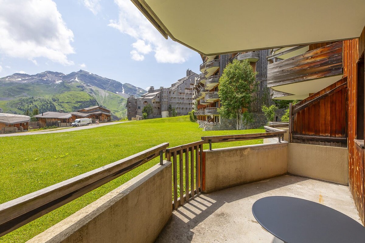 Balcony view over a vibrant green field, traditional wooden chalets, and a large ski resort building, with snowy mountains under a cloudy sky.