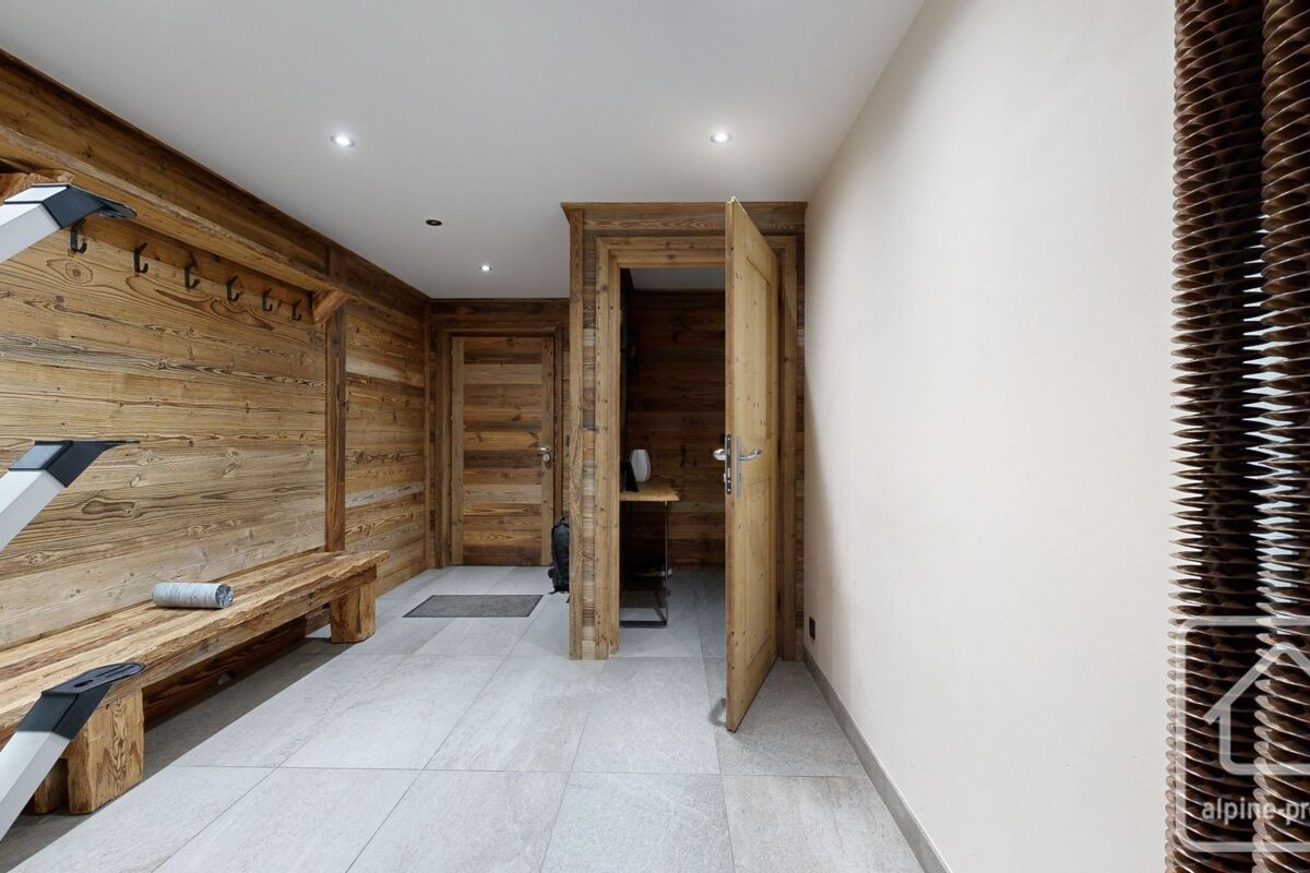 A rustic-modern room with wood paneling, light tiled floor, a wooden bench, coat hooks, and multiple doors, hinting at an alpine or spa setting.