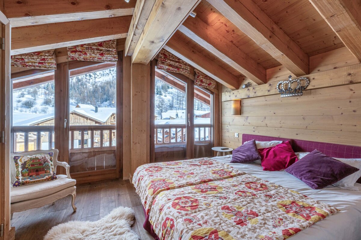 Cozy wooden cabin bedroom with exposed beams, a colorful bed, and large windows offering a snowy mountain view.