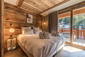 Cozy, wood-paneled bedroom with a plush bed, white linens, and slippers. A balcony door reveals a snowy forest and mountain view.