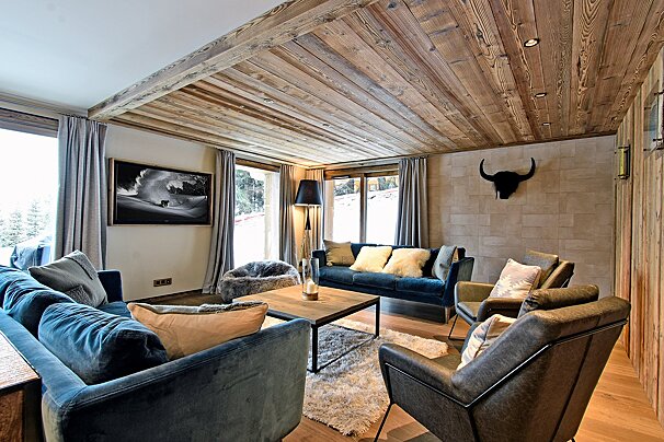Elegant chalet living room with rustic wood ceiling, plush blue sofas, leather armchairs, and a bold bull skull, overlooking a snowy landscape.