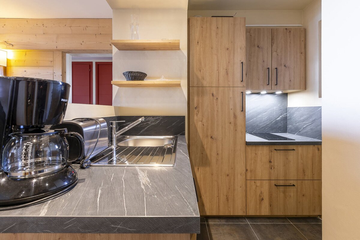 Modern kitchen with wood cabinets, dark marbled counters, coffee maker, toaster, and sink. Floating shelves and red-framed window visible.