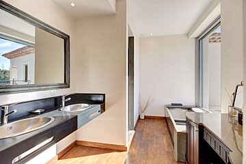 Bright, modern bathroom with a double vanity, large mirror, sleek bathtub, and wooden floor, illuminated by a big window.