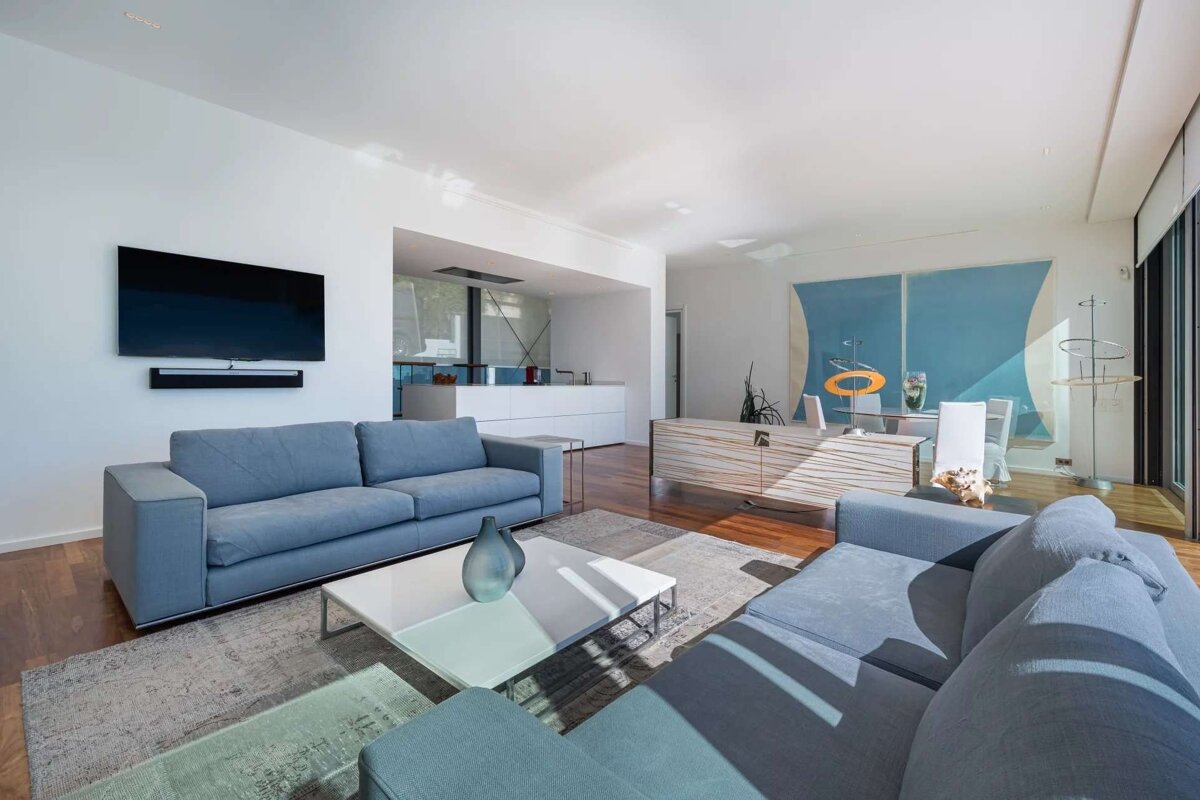 A bright, modern open-plan living room with blue sofas, a white coffee table, and a wall-mounted TV. A kitchen and dining area are visible.