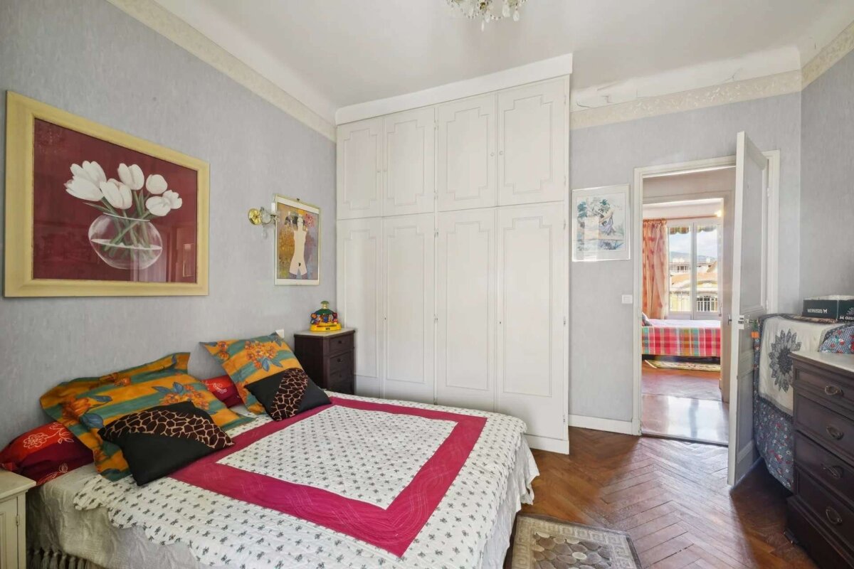 Cozy bedroom with a colorful bed, white wardrobe, hardwood floors, artwork, and a doorway leading to another sunlit room.