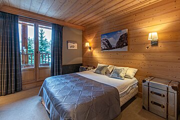 A cozy, chalet-style bedroom features warm wood paneling, a large bed with sheep pillows, wall sconces, and a balcony overlooking pine trees.