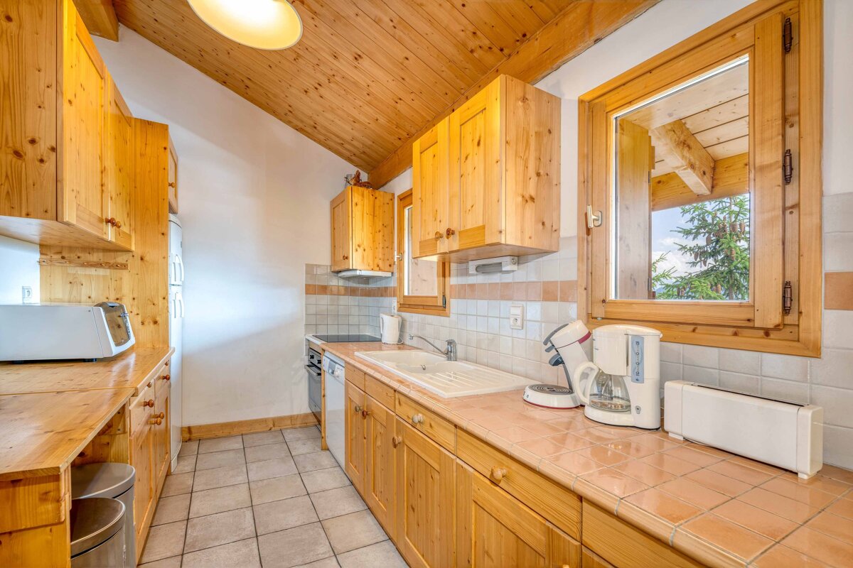 Cozy, rustic kitchen featuring wooden cabinets, a sloped wood ceiling, tiled counters, and appliances. A window offers a view of trees.