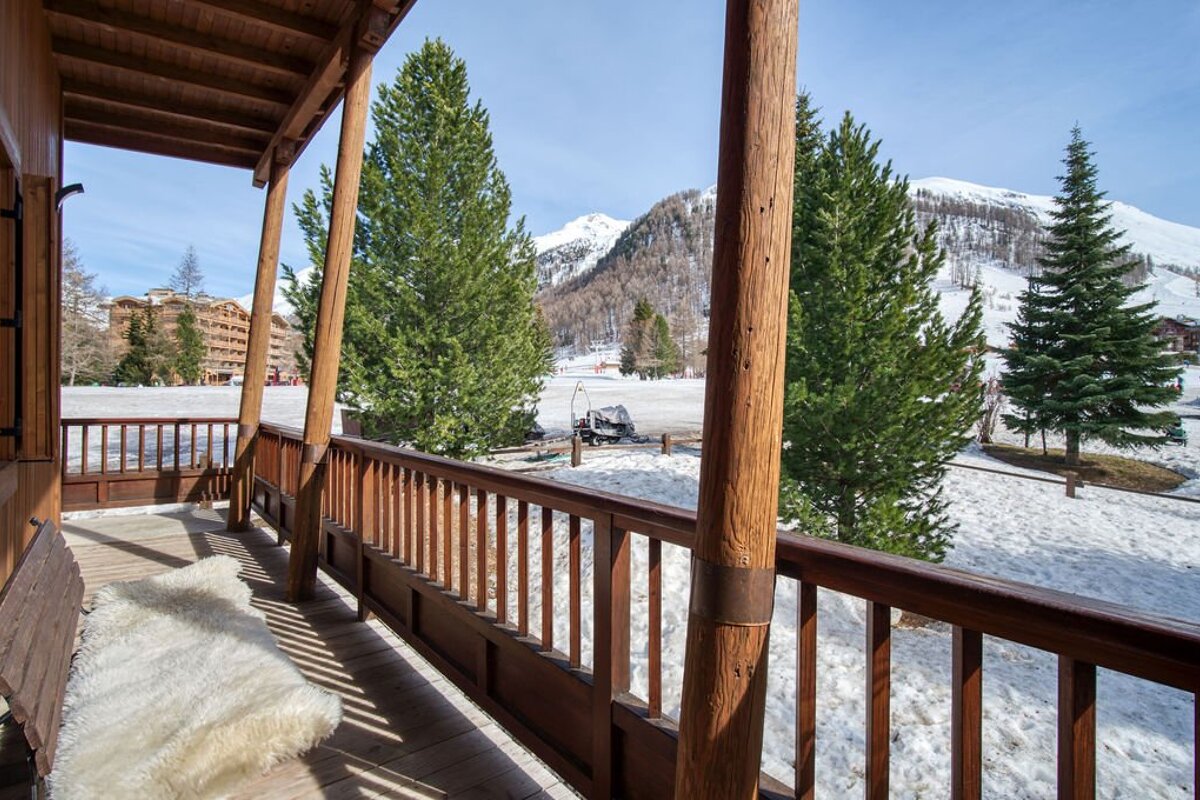 Cozy wooden balcony with a fluffy rug overlooks a snowy mountain ski resort valley, featuring pine trees, distant buildings, and a sunny sky.