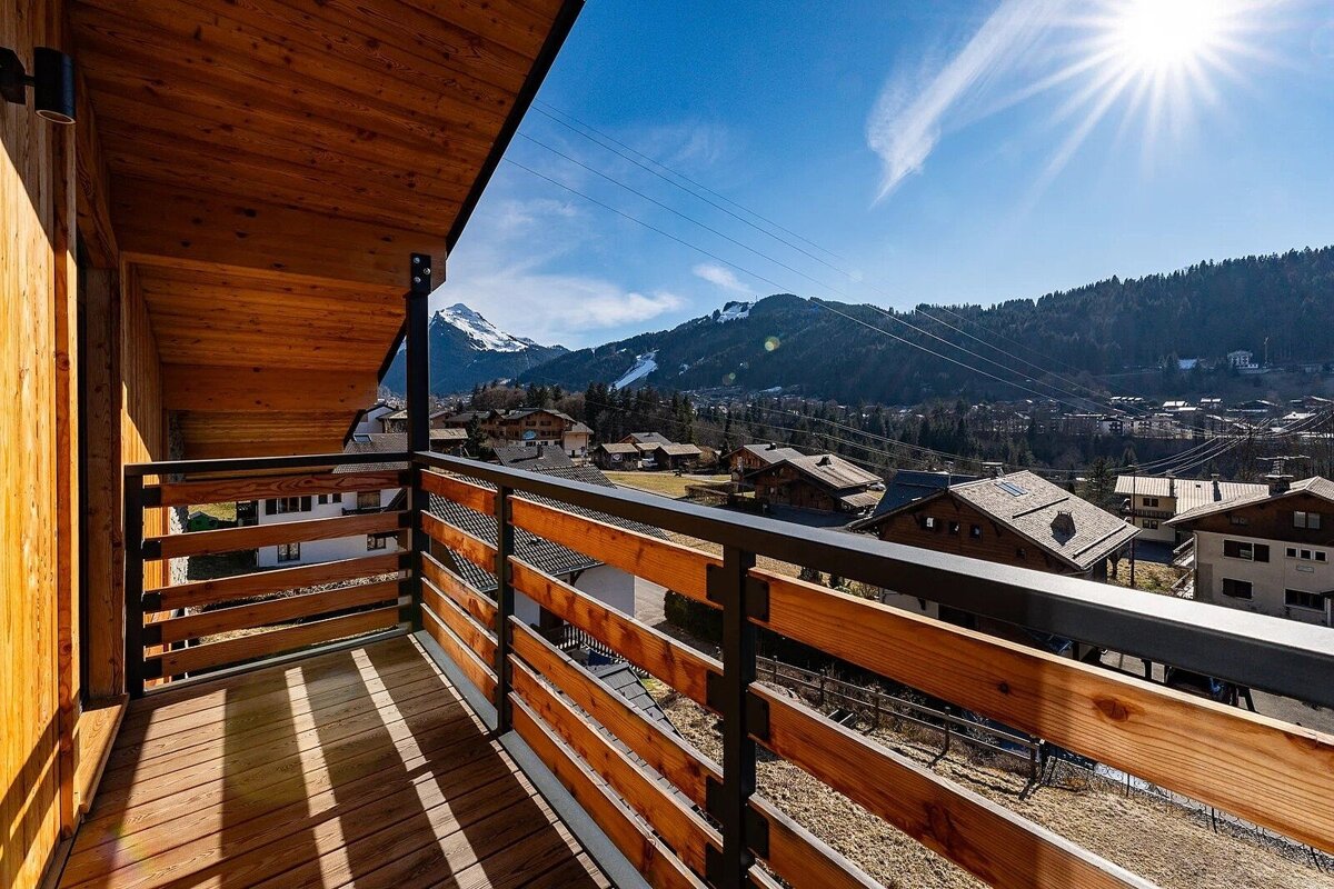 A wooden balcony offers a sunny view of an alpine village with snow-capped mountains and ski slopes under a clear blue sky.
