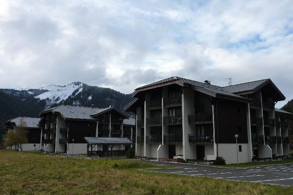 Chalet-style buildings with balconies stand in a grassy field against a backdrop of snow-capped mountains and a cloudy sky.