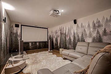 A modern home theater room features forest mural wallpaper, a projector screen, gray sectional sofa, and a white fluffy rug on a wooden floor.