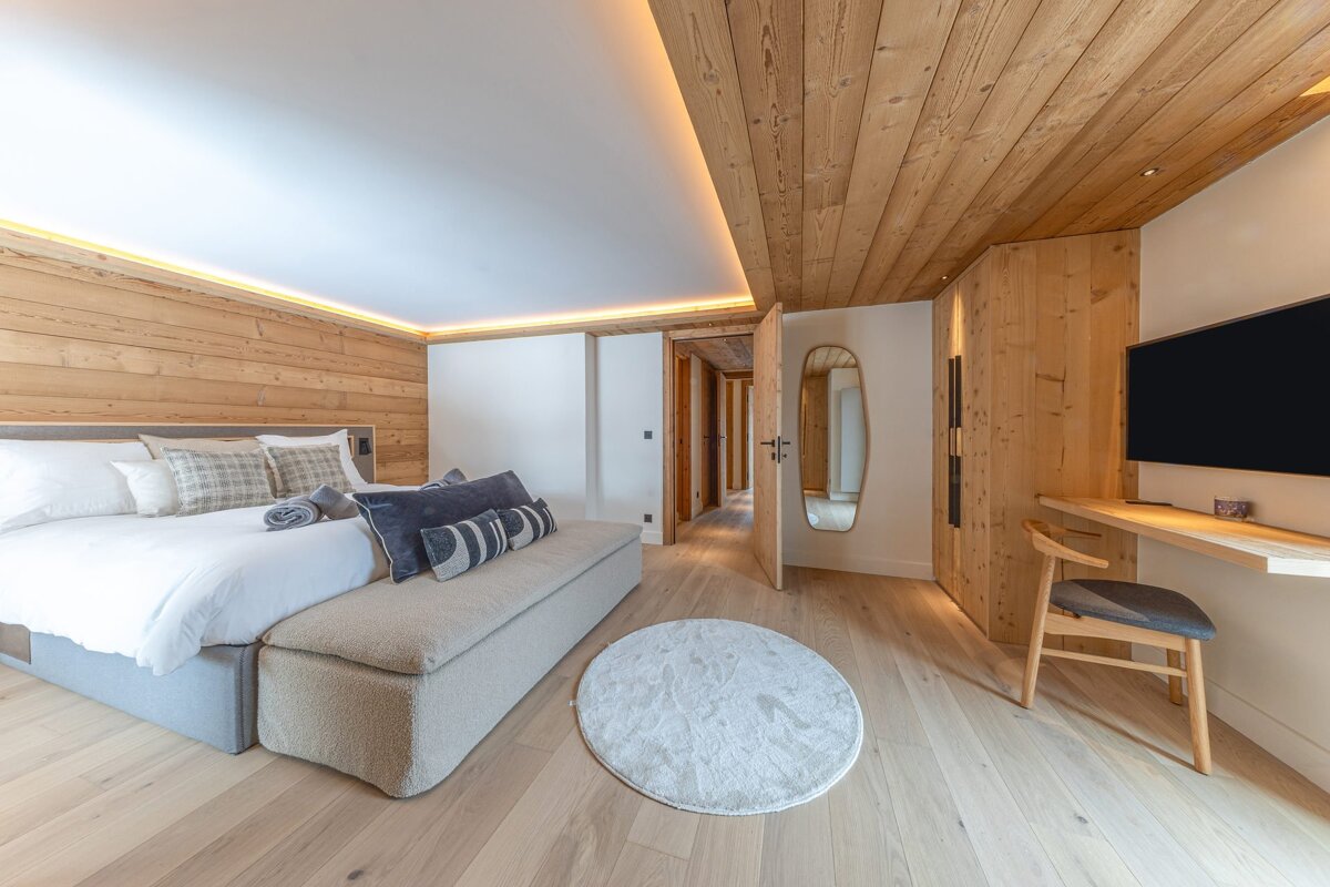 A chic bedroom features natural wood paneled walls/ceiling, a large bed, ambient lighting, light hardwood floors, and a desk with a TV.