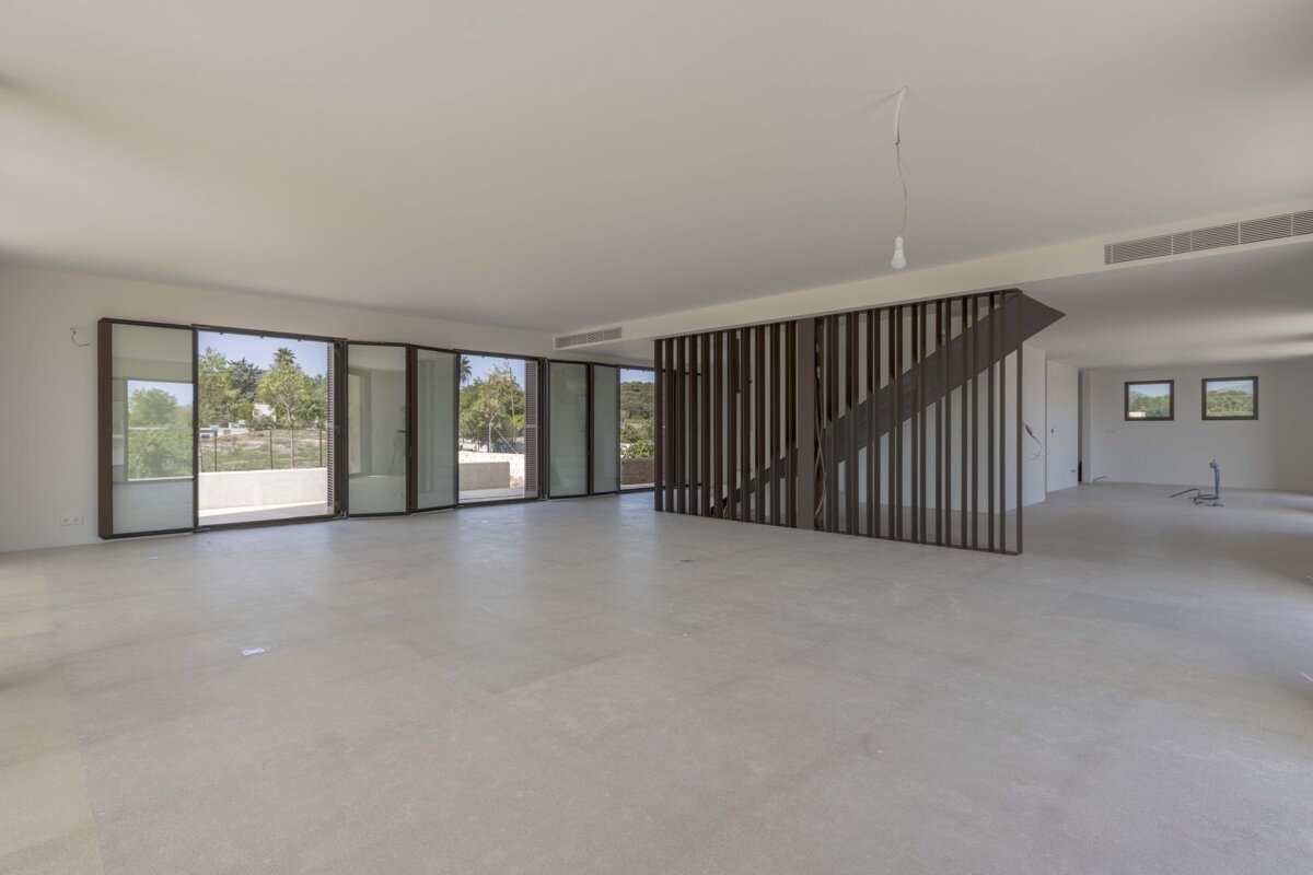 Large, empty modern room with light floors, expansive glass doors overlooking a green landscape, and a stylish slatted wooden staircase.