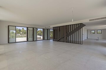 Large, empty modern room with light floors, expansive glass doors overlooking a green landscape, and a stylish slatted wooden staircase.