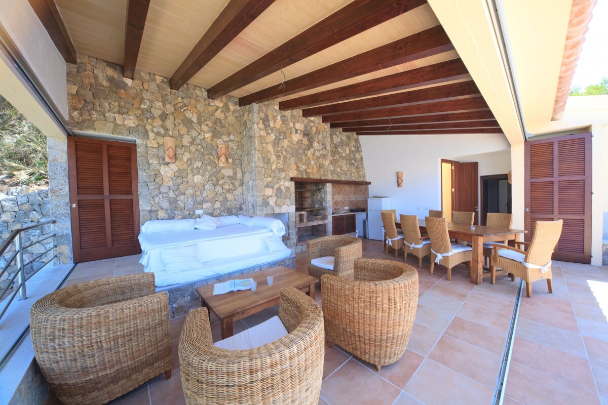 Spacious stone-walled outdoor patio with wooden beams, featuring a lounge area with wicker furniture, a daybed, an outdoor kitchen, and a dining table.