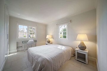 Bright, minimalist bedroom with white walls, light floor tiles, a bed, two windows, two lamps, and a small desk with a clear chair.