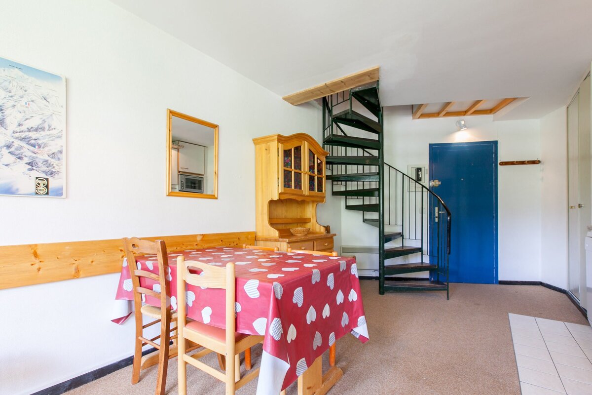 A room with a dining table, heart tablecloth, wooden hutch, black spiral staircase, and a blue door. Ski map and mirror on white walls.