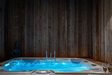 A luxurious hot tub with glowing blue water and jets, nestled against dark wooden walls with a Buddha statue and champagne flutes nearby.