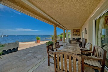 A covered patio with wooden dining furniture offers a stunning view of the blue sea with boats, creating a serene outdoor dining space.