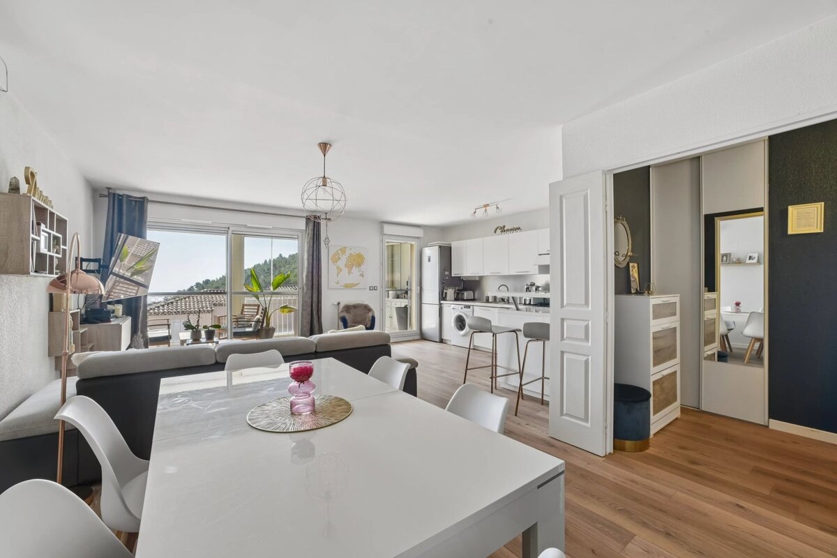 Bright, open-concept apartment featuring a white dining table, a living area with balcony access, and a modern kitchen.