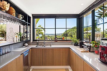 Modern kitchen featuring light wood cabinets, gray counters, and large black-framed windows with a beautiful landscape view.