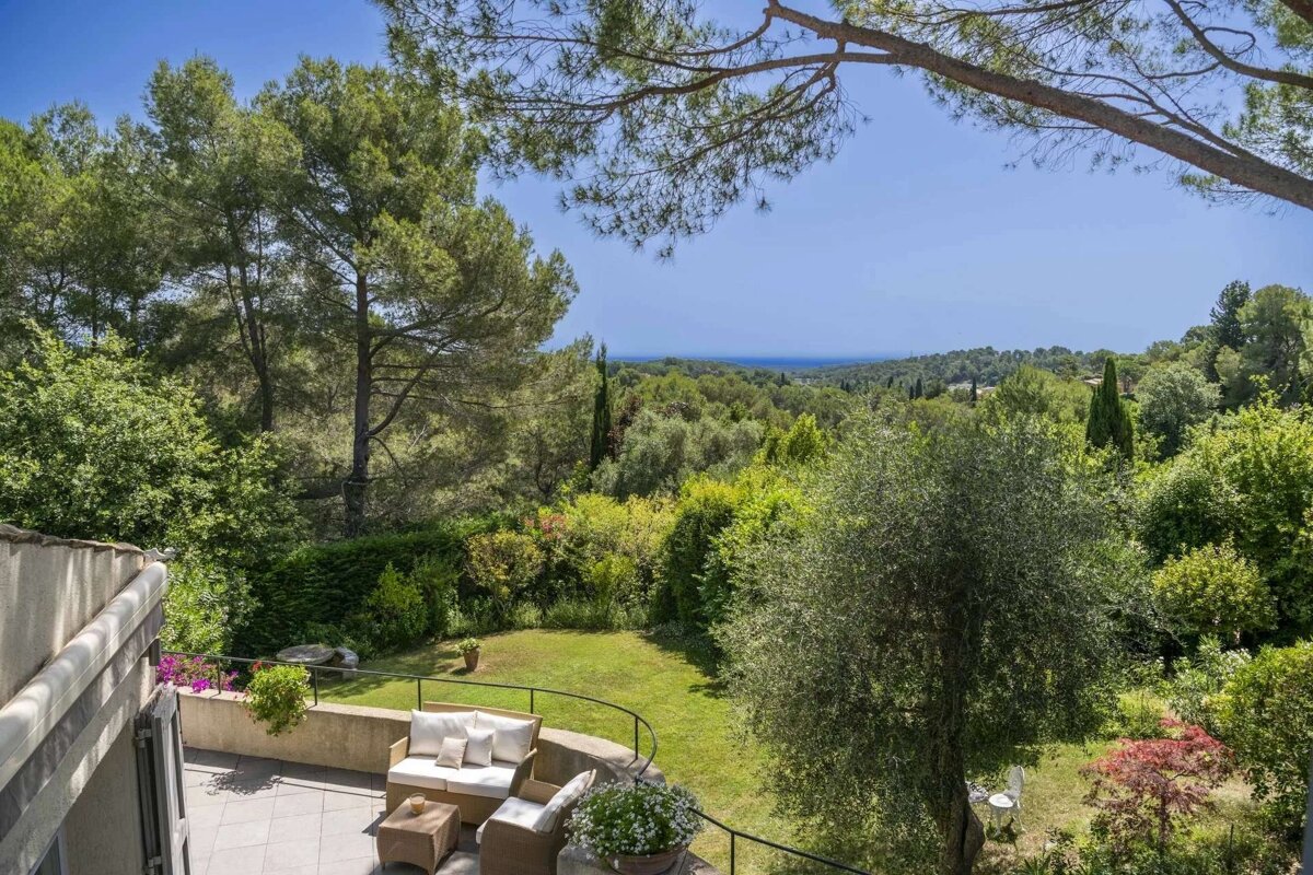 A sunny patio with wicker furniture overlooks a lush garden, dense forest, and distant blue sea, creating a tranquil Mediterranean landscape.