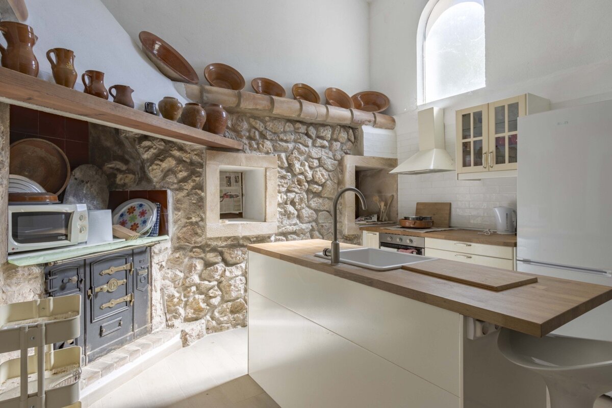 A rustic kitchen with exposed stone walls, traditional oven, and terracotta decor, contrasted by a modern white island with a sink and contemporary white appliances.