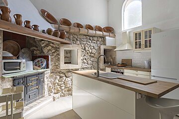 A rustic kitchen with exposed stone walls, traditional oven, and terracotta decor, contrasted by a modern white island with a sink and contemporary white appliances.