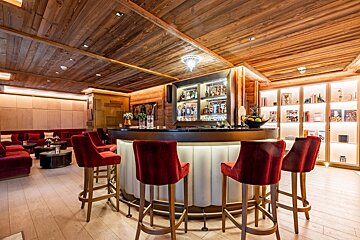 An elegant bar and lounge with rich wooden ceilings, a circular bar with red velvet stools, and cozy red sofas for seating.