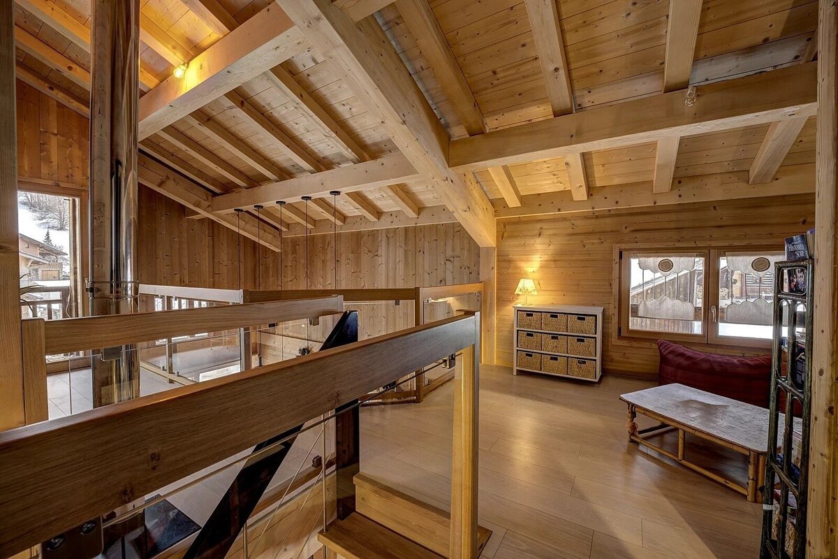 Cozy, rustic wooden chalet interior with high ceilings, exposed beams, glass railings, and natural light from large windows showing a snowy view.