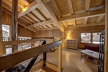 Cozy, rustic wooden chalet interior with high ceilings, exposed beams, glass railings, and natural light from large windows showing a snowy view.