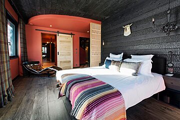 Eclectic bedroom with dark charred wood & vibrant coral walls. Features a white bed with colorful throw, rustic barn door, and modern chaise lounge.