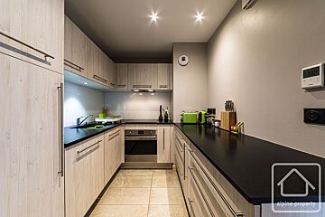 A narrow kitchen with light wood cabinets, black countertops, and beige tiled flooring.