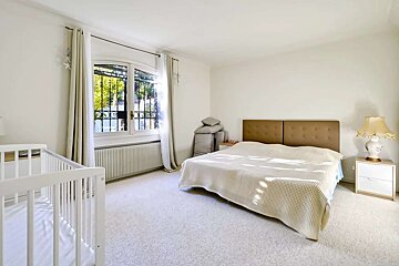 Bright, carpeted bedroom with a large bed, white crib, and sunlit window. Features light walls, brown headboard, and a classic lamp.