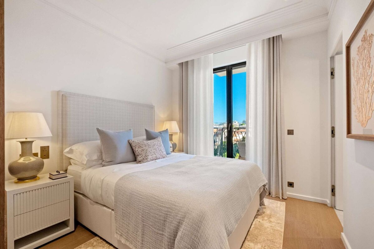 Bright, modern bedroom featuring a bed with neutral linens, nightstands, lamps, and a balcony door overlooking a sunny sky and cityscape.