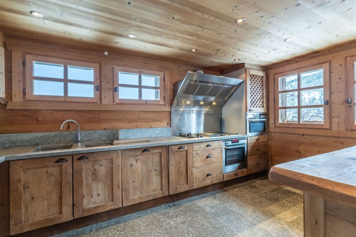 A rustic wooden kitchen with granite counters, stainless steel appliances, and multiple windows. Features a stone floor and a warm, natural aesthetic.