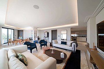A modern open-plan living room with light sofas, blue accent chairs, a dining area, and a visible kitchen, featuring stylish decor and ambient lighting.