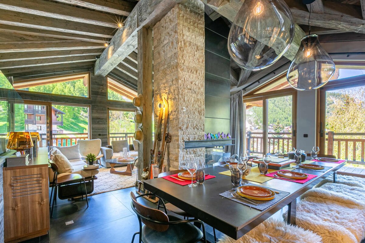 A luxurious modern chalet interior: open-plan dining/living, exposed wooden beams, stone fireplace, and large windows framing stunning mountain views.