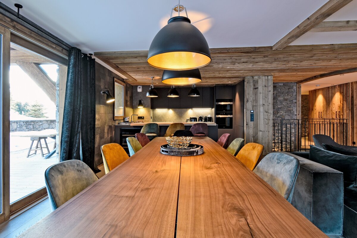 Cozy, modern chalet dining area with a long wooden table, multi-colored chairs, open kitchen, and warm lighting, looking out to a snowy landscape.