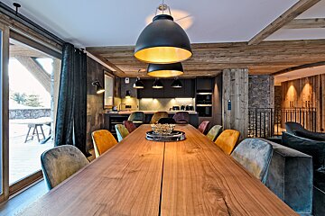 Cozy, modern chalet dining area with a long wooden table, multi-colored chairs, open kitchen, and warm lighting, looking out to a snowy landscape.