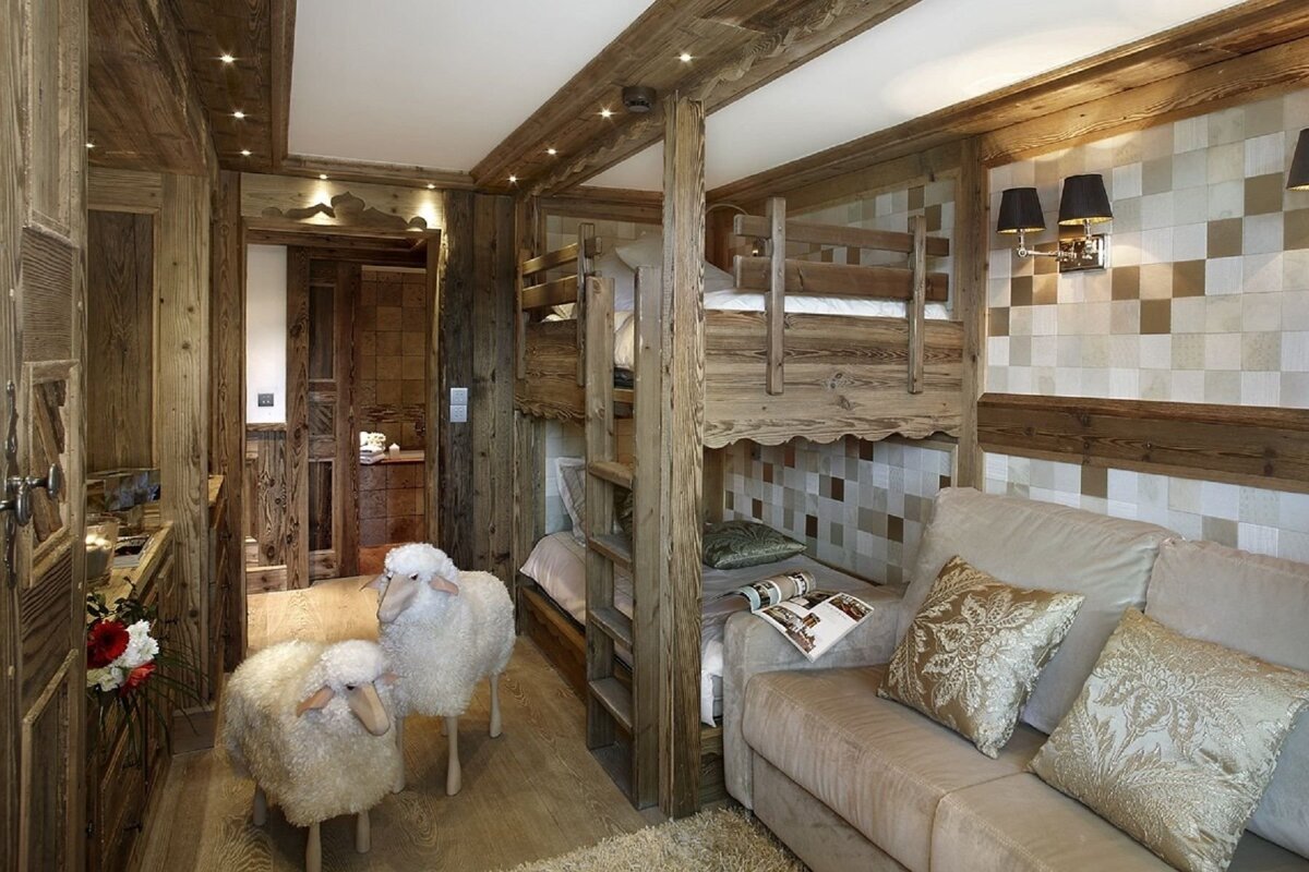 A cozy, rustic wooden room featuring bunk beds, a sofa, and two fluffy sheep statues. Warm lighting and patterned walls complete the chalet style.