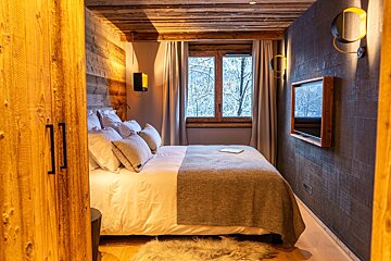 A cozy, rustic bedroom featuring a bed, TV, warm lighting, and a window revealing a snowy winter view. Wood walls and ceiling create a warm ambiance.