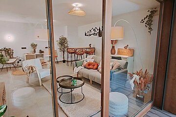Cozy boho-style living room with a grey sofa, warm lighting, and natural accents, viewed through a glass sliding door.