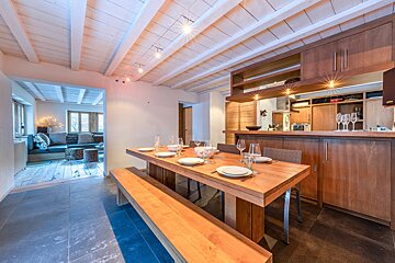 A cozy, open-plan dining area with a wooden table, benches, and a modern kitchen, leading into a relaxed living room with white beamed ceilings.