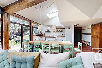 Bright open-concept living area with a large dining table, green chairs, a kitchen, and views of a lush outdoor area.