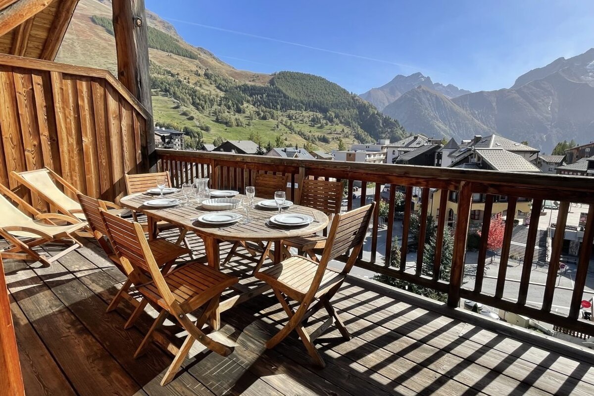 Wooden balcony with a set dining table overlooking a stunning mountain village and alpine landscape.